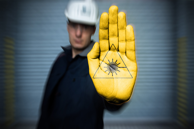 Mann mit Bauhelm hält seine gelb gefärbte Handfläche mit dem Gefahrenzeichen „Laserstrahlung“ in die Kamera.