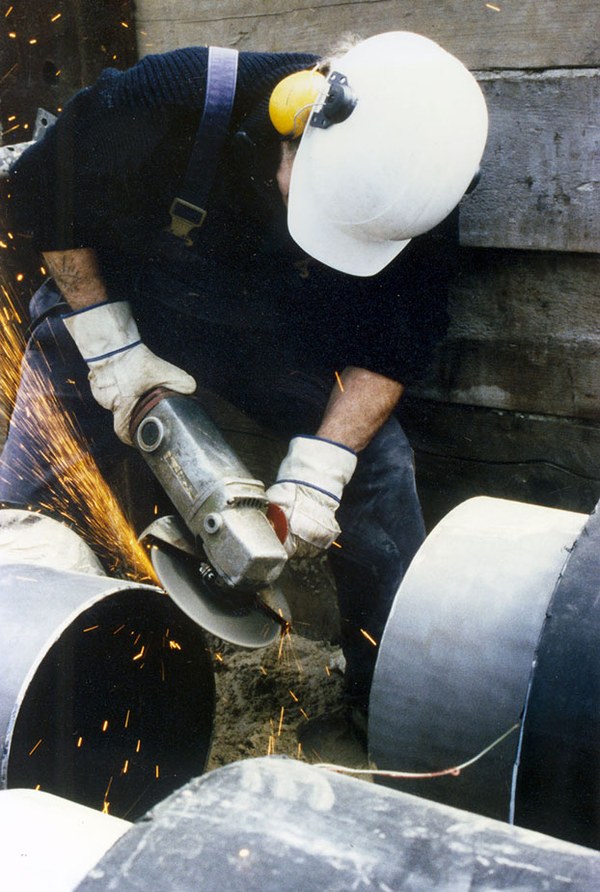 Arbeiter, der Helm, Gehörschutz und Sicherheitshandschuhe trägt, bei Arbeiten mit dem Winkelschleifer in einem engen Rohrgraben