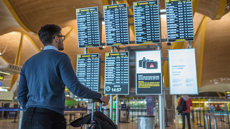 Passagier mit Reisetrolley schaut auf eine Reihe von Displays mit Abflugzeiten