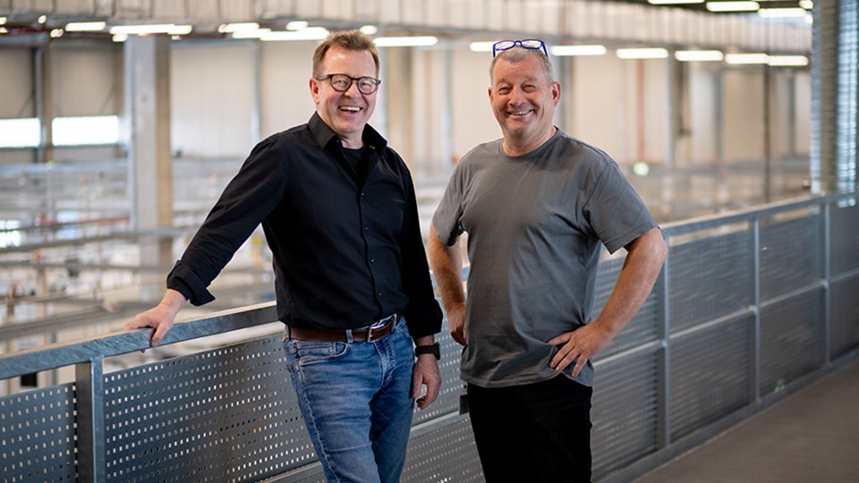 Ulrich Schwab und Andreas Neugebauer stehen nebeneinander auf der Empore einer Produktionshalle und lachen in die Kamera.