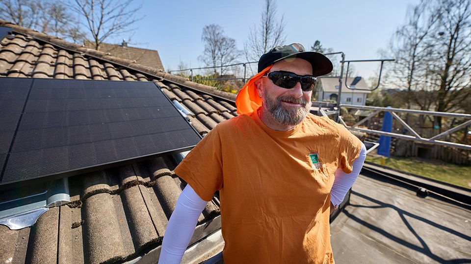 Mitarbeiter von Graf Solar auf einem eingerüsteten Dach mit Solarpanelen, er trägt Sonnenbrille, eine Schirmmütze mit Nackenschutz und unter dem orangefarbenen T-Shirt weiße Armstulpen