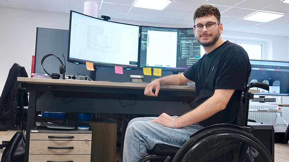 Serhat Yüksel im Rollstuhl an seinem Arbeitsplatz mit zwei Monitoren, im Hintergrund des Büroraums ein Bildschirm für Präsentationen