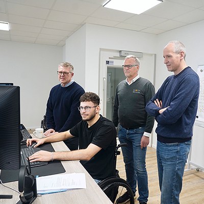 Serhat Yüksel an seinem Bildschirmarbeitsplatz, drei Männer stehen hinter ihm und schauen gemeinsam mit ihm auf den Monitor