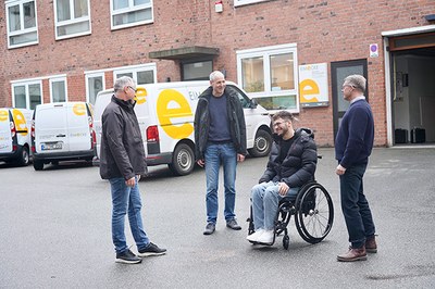 Michael Renner, Andreas Abel, Serhat Yüksel im Rollstuhl und Mathias Wolf auf Firmengelände, im Hintergrund Firmenfahrzeuge