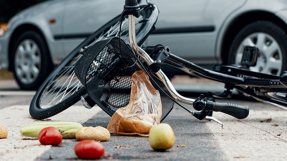 Ein Fahrrad, Obst und andere Einkäufe liegen nach einem Unfall auf der Straße, im Hintergrund ein silberner Pkw