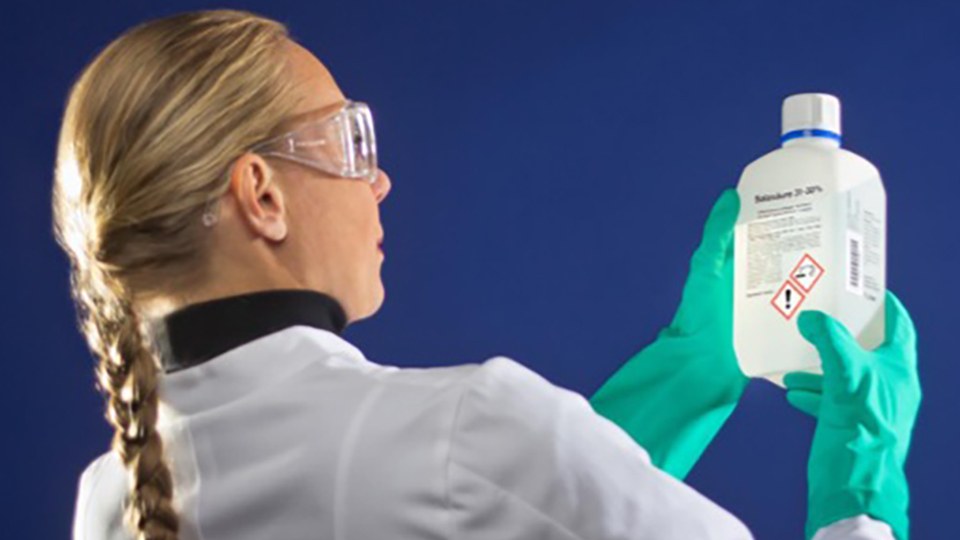 Labormitarbeiterin mit Schutzbrille, Kittel und Sicherheitshandschuhen schaut auf das Etikett einer Chemikalienflasche.