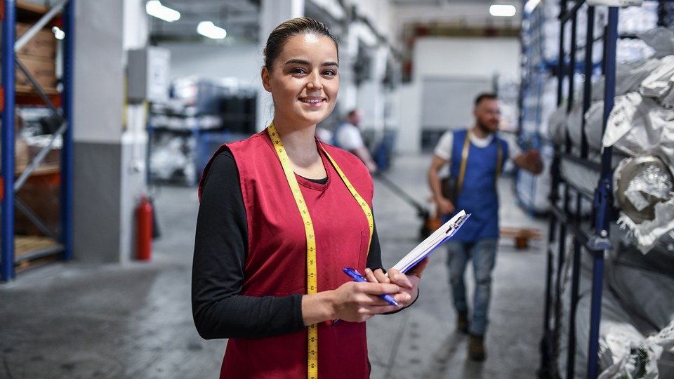Auszubildende in der Lagerhalle einer Textilfabrik. Sie lächelt in die Kamera, hält ein Clipboard in der Hand und trägt ein Maßband am Hals. Im Hintergrund ein Kollege.