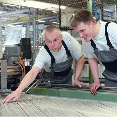 Ausbilder und Auszubildender beugen sich über eine Webmaschine und begutachten das Gewebe.