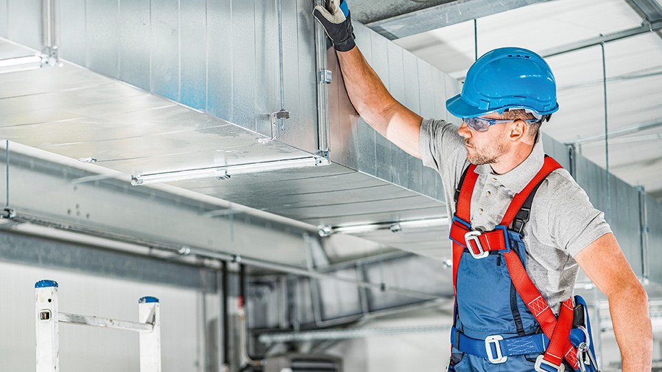 Monteur prüft in einer Fabrik die Lüftungskanäle einer raumlufttechnischen Anlage. Er trägt Helm, Schutzbrille, Arbeitshandschuhe und ist mit einem Geschirr gesichert.