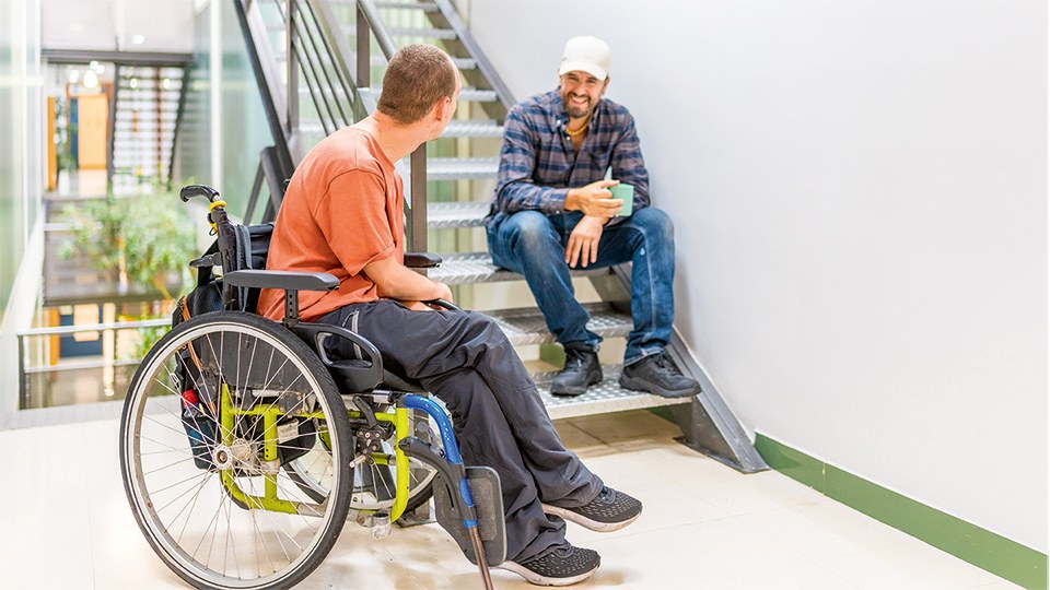 Zwei Kollegen unterhalten sich in der Pause. Einer von beiden sitzt in einem Rollstuhl, der andere sitzt auf den Stufen einer Metalltreppe ein einem Industriegebäude.