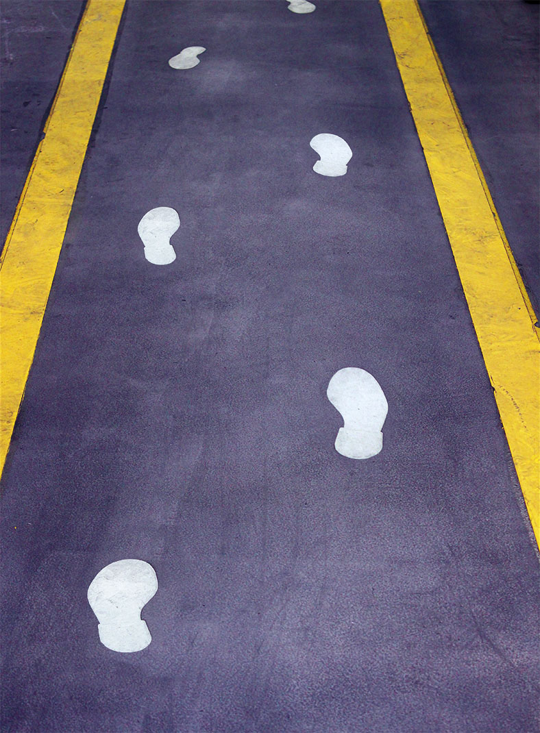 Das Bild zeigt eine Werkshalle mit grauem Fußboden. Der zu benutzende Fluchtweg ist markiert durch gelbe Randstreifen und mit weißen Schuhabdrücken versehen. 