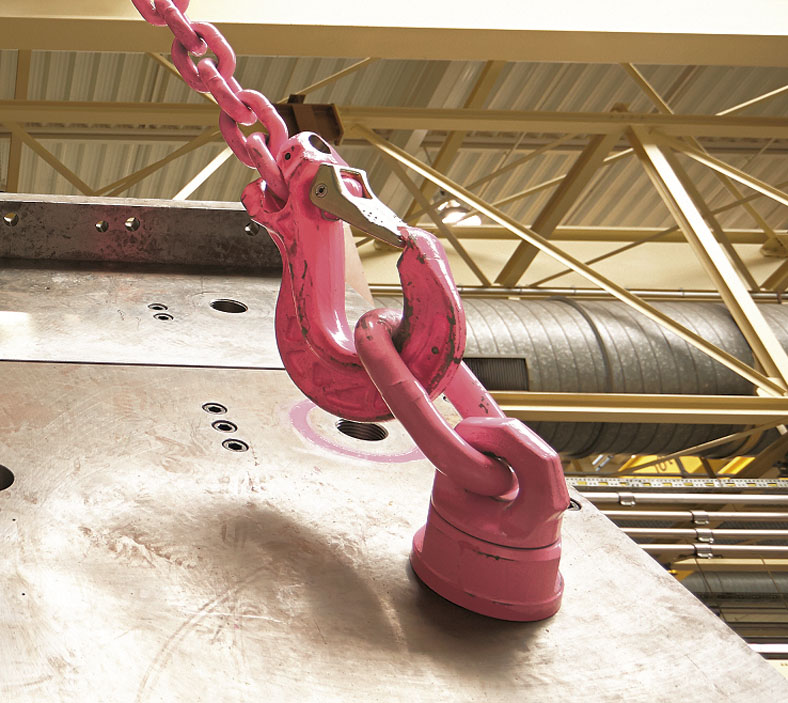 Das Bild zeigt in einer Fabrikhalle einen großen roten Lasthaken an einer Kette, der in die rote Metallschlaufe eines Lastbockes eingehängt ist, welcher an einer schrägen Metallplatte angebracht ist.