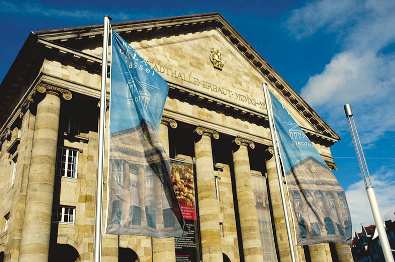 Das Bild zeigt die Front des Kongress Palais in Kassel mit Säulenportikus vor einem blauen Himmel. Im Vordergrund zwei Stangen mit wehenden blauen Fahnen, die Abbildungen der Stadthalle zeigen.