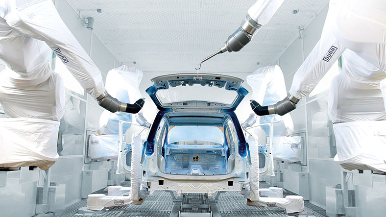 Das Foto ziegt den hinteren Teil einer Autokarosserie mit geöffneter Heckklappe, die in einer Werkshalle von Roboterarmen montiert wird. Die Roboter sind mit weißen Stoffen verkleidet.