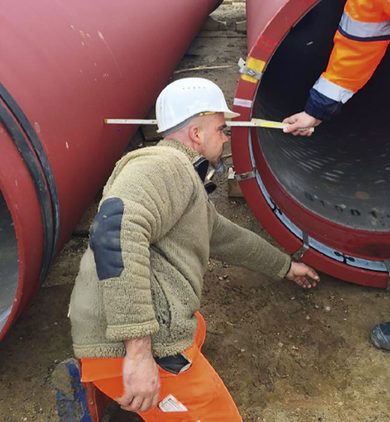 Auf dem Bild kniet ein Arbeiter mit weißem Schutzhelm vor zwei roten Abwasserrohren, daneben sieht man den Arm eines anderen Arbeiters, der mit einem Meterstab den Abstand zwischen zwei Rohren misst.