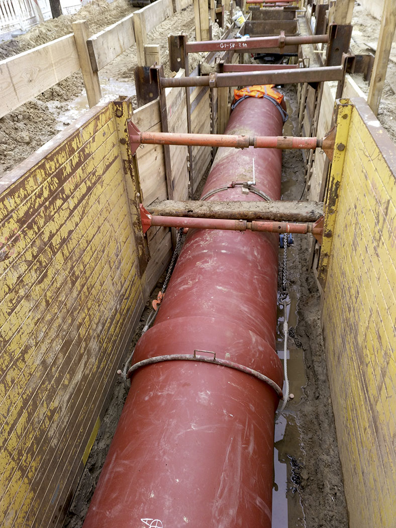 Das Bild zeigt einen offenen Graben mit einer roten Abwasserrohrleitung, die durch Kettenzüge zusammengezogen ist. Der Graben ist durch Montagestangen abgestützt.