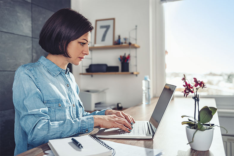 Das Foto zeigt eine Frau im Profil mit dunklem Pagenschnitt und blauer Bluse. Sie sitzt an zuhause an einem Schreibtisch und tippt auf einer Laptop-Tastatur, rechts im Bild steht eine blühende Topfpflanze auf dem Tisch. Im Hintergrund sieht man ein Wandregal und ein Fenster mit Blick auf blauen Himmel.