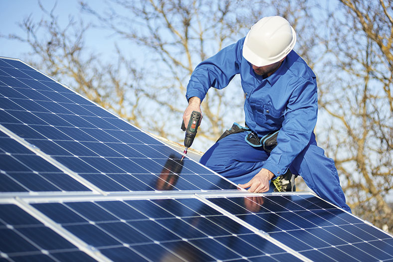 Auf dem Foto sieht man einen Arbeiter in Blaumann und weißem Schutzhelm auf einem Dach. Er montiert Solarzellen mit einem Akkuschrauber. Im Hintergrund Äste eines Baumes und blauer Himmel.