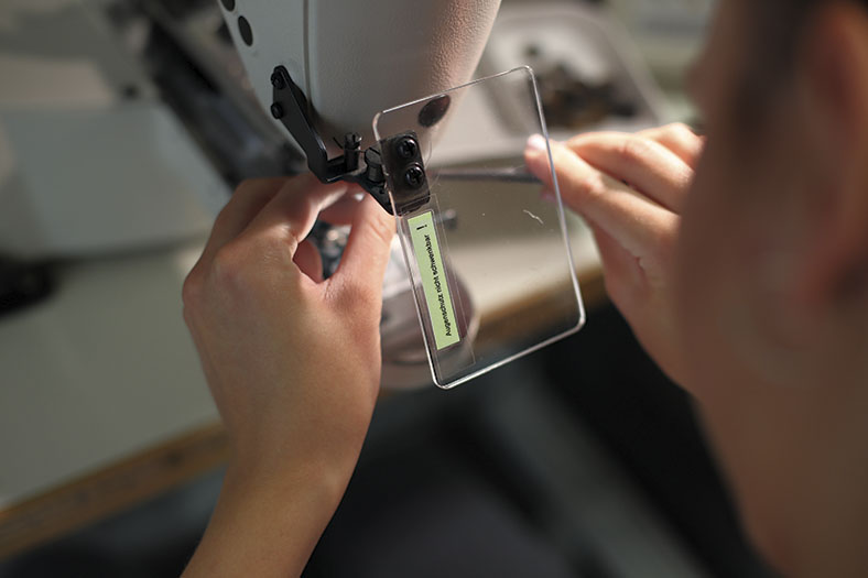 Das Foto zeigt, wie Frauenhände etwas an der Nadelhalterung einer Nähmaschine montieren. Vor dem Nadelbereich ist ein transparenter Schuztschild an der Maschine angebracht.