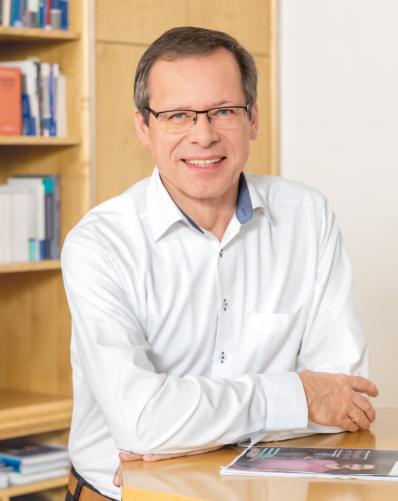 Porträtfoto von Johannes Tichi, Vorsitzender der Geschäftsführung der BG ETEM. Er hat kurze Haare und eine Brille. Er trägt ein helles Hemd und stützt beide Arme auf einen Stehtisch, auf der die neue Ausgabe des etem-Magazins liegt.