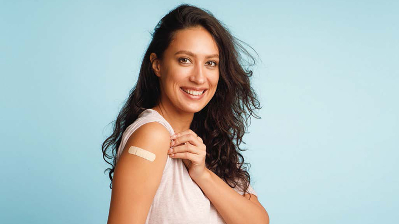 Junge Frau mit langen dunklen Haaren in einem weißen Oberteil vor hellblauem Hintergrund. Sie hält ihren Ärmel hoch, um ein Impfpflaster zu zeigen und lächelt dabei in die Kamera.