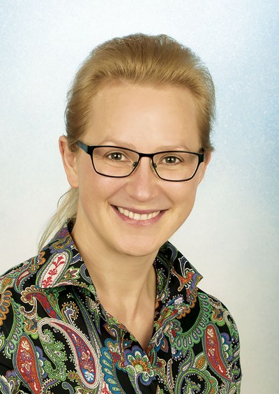 Porträt von Jenny Blumenthal, einer Außendienstmitarbeiterin und Aufsichtsperson der BG ETEM. Sie trägt lange blonde Haare zu einem Pferdeschwanz gebunden, eine schwarz-gerahmte Brille, eine bunte Bluse und lächelt in die Kamera.