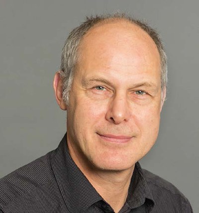 Porträtfoto von Dr. Just Mields, Arbeitspsychologe bei der BG ETEM. Herr Mields hat kurze graue Haare, trägt ein dunkles Hemd und lächelt. 