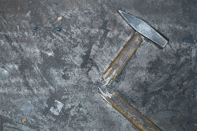 Motiv Arbeitsunfall: Ein Hammer mit zerbrochenem Holzstiel liegt auf einem grauen Untergrund.