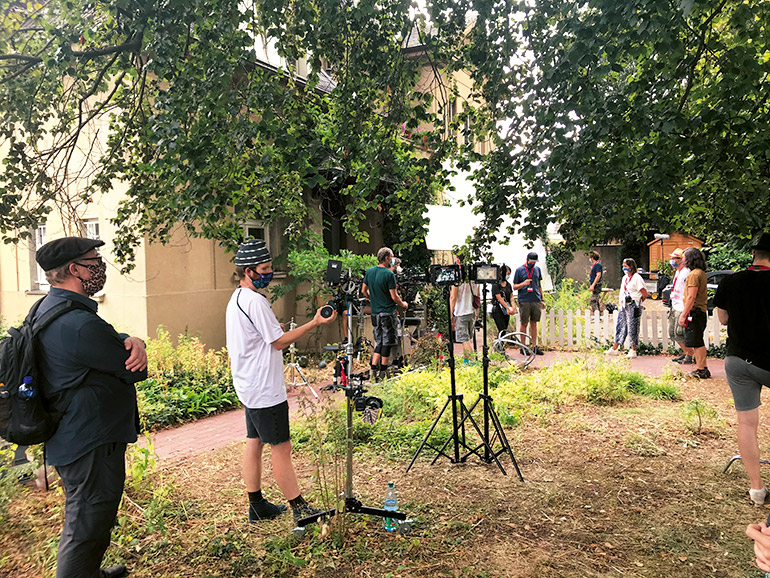 Sicherheit bei Filmproduktionen: Szene an einem Filmset. Mehrere Personen stehen in einem Garten vor einem Haus, um sie herum Filmausrüstung wie Lampen und Kameras.