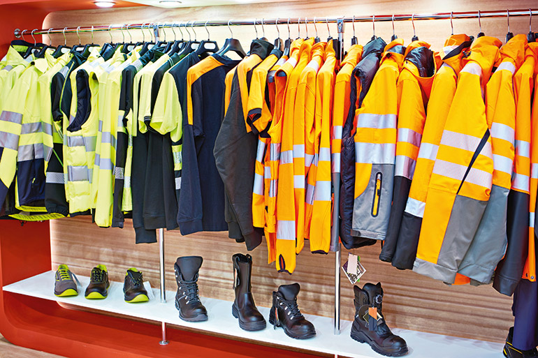 Kleidung Arbeitsschutz: Kleiderstange mit gelben und orangen Sicherheitswesten, -jacken- und Shirts, darunter auf einem Regalbrett mehrere Sicherheitsschuh-Modelle.