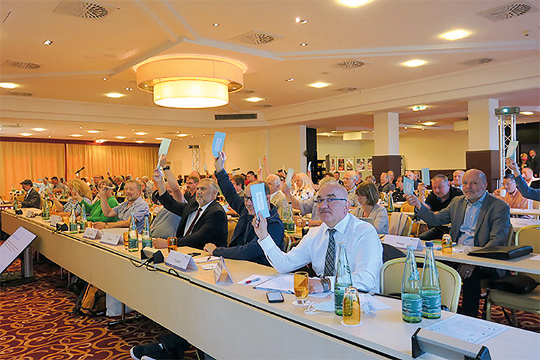 Vertreterversammlung der BG ETEM in Heidelberg, die Anwesenden sitzen in einem Hotelsaal an Tischreihen, Abstimmung der Anwesenden per Handheben zur Unternehmerpflichtversicherung.