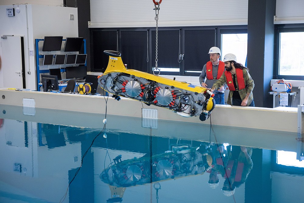 Forschung und Arbeitsschutz: Felix Bernhard und Dr. Bilal Wehbe bewachen den gelben Roboter „Flatfish“ beim Hinabsinken in ein Unterwasser-Trainingsbecken. Sie tragen beide Bauhelm und Rettungsweste.