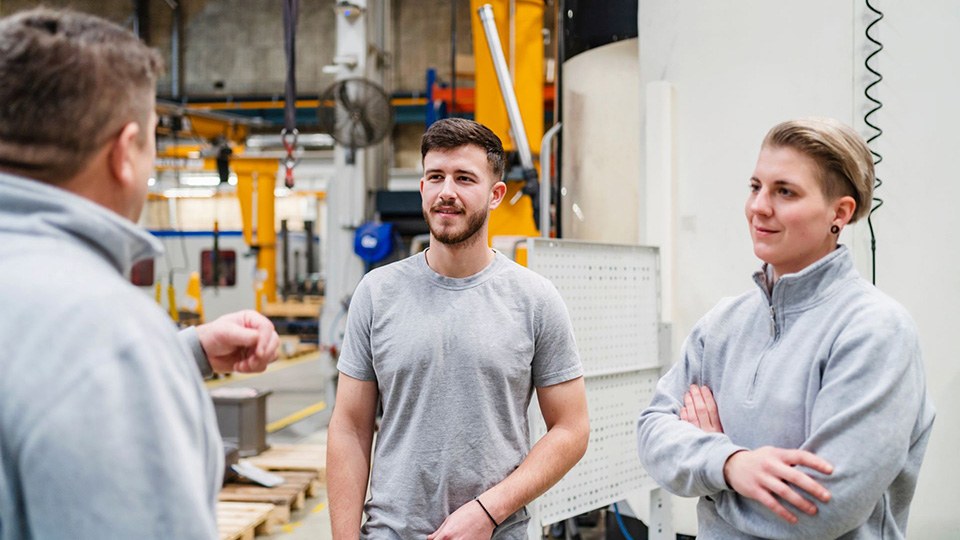 Zwei Angestellte – ein Mann und eine Frau –  in einer Fabrik hören einem Kollegen zu