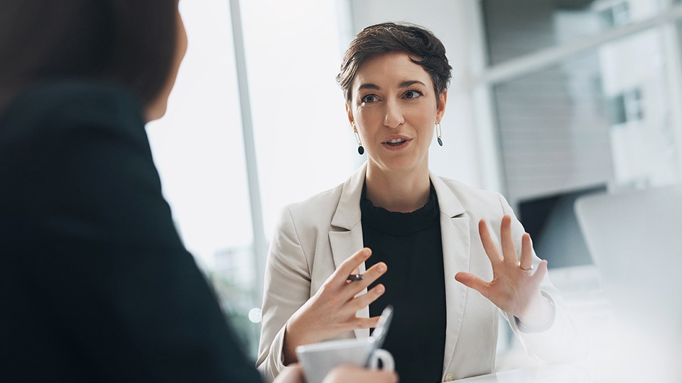 Zwei Frauen an einem Tisch in Bürosituation im Gespräch, die eine redet und gestikuliert dabei