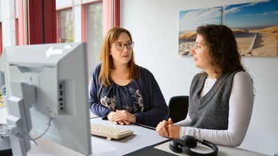Zwei Frauen stehen im Büro zusammen an einem höhenverstellbaren Schreibtisch mit Computerarbeitsplatz und unterhalten sich