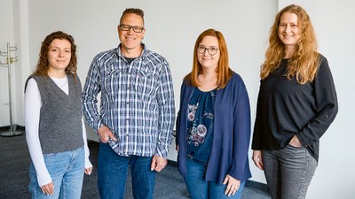 Drei Frauen und ein Mann stehen in einem Besprechungsraum nebeneinander und lächeln in die Kamera.