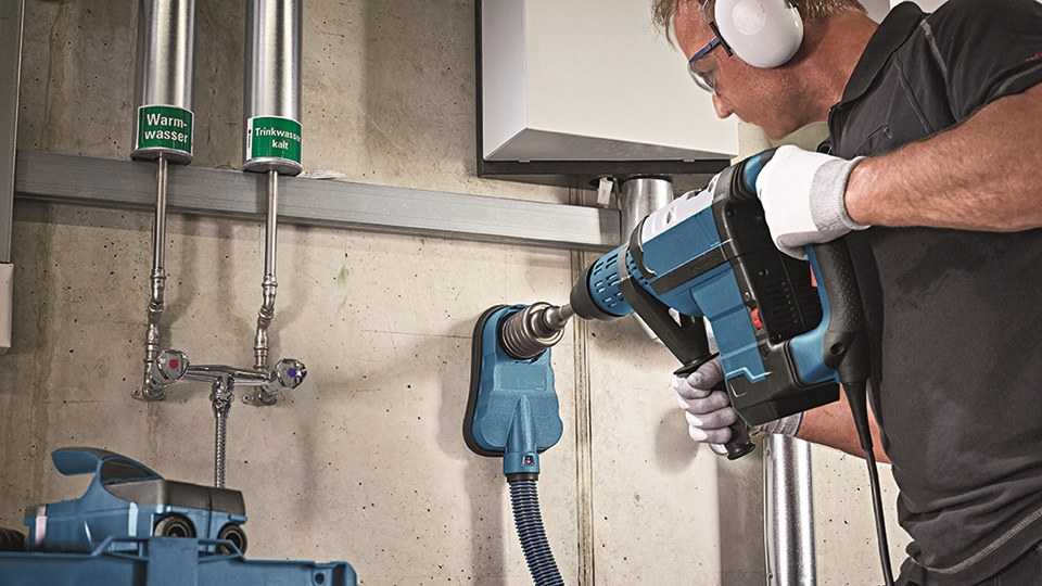 Arbeiter mit Arbeitshandschuhen bohrt mit einer Hohlbohrkrone ein Loch in eine Betonwand; an den Bohrhammer ist eine Absaugung angeschlossen, die den Staub über einen Schlauch abführt.