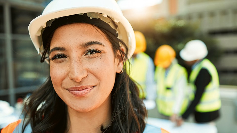 Junge Frau mit Schutzhelm und Sicherheitsweste auf einer Baustelle, sie lächelt in die Kamera, hinter ihr unscharf drei Kollegen