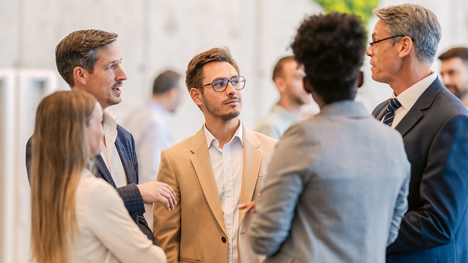 Eine Gruppe von drei Männern und zwei Frauen in legerer Businesskleidung stehen auf einer Veranstaltung zusammen und diskutieren.