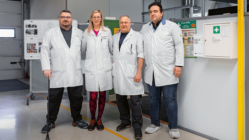 Drei Männer und eine Frau stehen in einer Fabrikhalle Arm in Arm nebeneinander und blicken in die Kamera. Alle tragen weiße Arbeitsschutzkittel. Ganz rechts hängt ein Erste-Hilfe-Schrank.