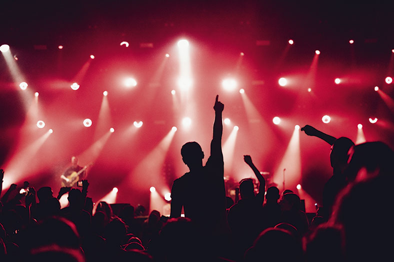 Auf dem Bild ist im Vordergrund eine Menschenmenge im Schatten zu sehen, die in den Hintergrund auf eine Bühne schauen, mehrere Personen strecken Arme in die Höhe. Im rot beleuchteten Hintergrund sieht man eine Bühne in rotes Licht getaucht sowie weiße Lichtkegel von mehreren Scheinwerfern. Auf der Bühne ist ein Mann mit einer Gitarre zu erkennen.