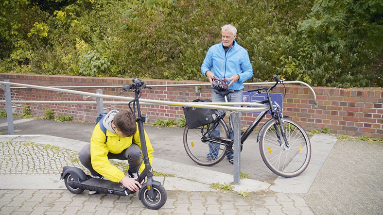 Ein Junge untersucht einen E-Scooter. Ein älterer Mann steht mit einem Fahrrad daneben.