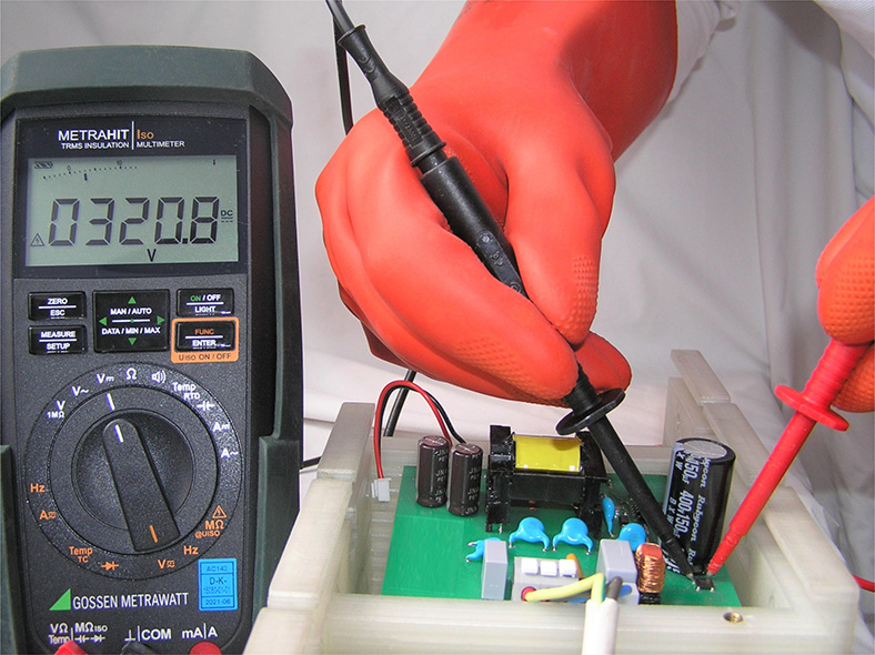 Elektrische Gefährdungen: Messen der Kondensatorspannung an einem unter Spannung stehenden Bauteil, links daneben Messgerät mit Spannungsanzeige.