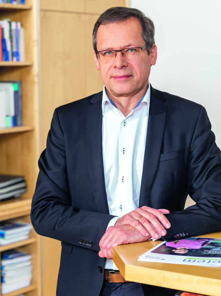 Porträtfoto von Johannes Tichi, Vorsitzender der Geschäftsführung der BG ETEM. Er hat kurze Haare und eine Brille und trägt eine Anzugjacke und ein weißes Hemd. Er hat den Arm auf einen Stehtisch gelegt, auf dem eine Ausgabe des etem-Magazins liegt.