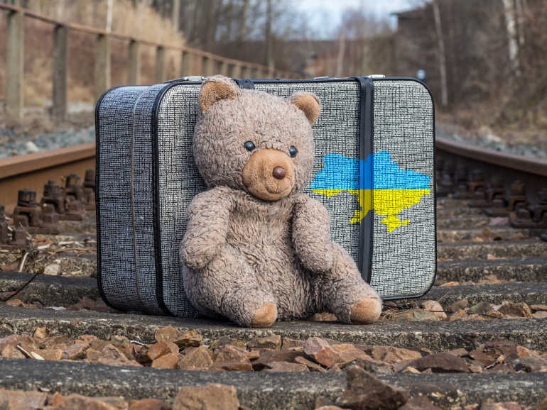 Ein Stoff-Teddybär sitzt angelehnt an einen grauen Koffer mit dem Umriss der Ukraine in blau und gelb auf den Schwellen einer Bahnlinie.