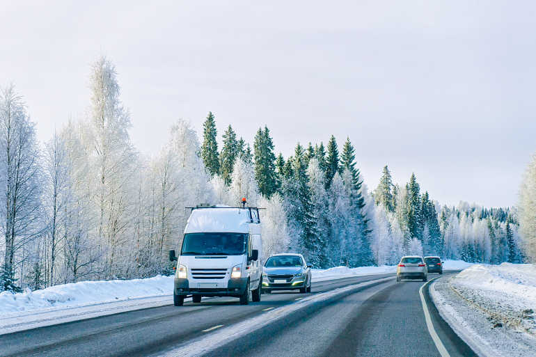 Drei Autos und ein Transporter fahren auf einer Straße, auf der etwas Schnee liegt. Die Straße ist. von schneebedeckten Bäumen gesäumt.