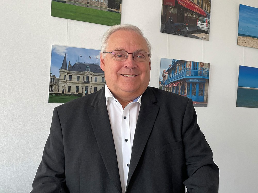 Entgeltmeldung: Markus Hellmann, Leiter Beitragsprüfung der BG ETEM, sitzt vor einer Wand mit Landschaftsfotografien. Er trägt einen dunklen Anzug und ein weißes Hemd, hat graue Haare und eine Brille.