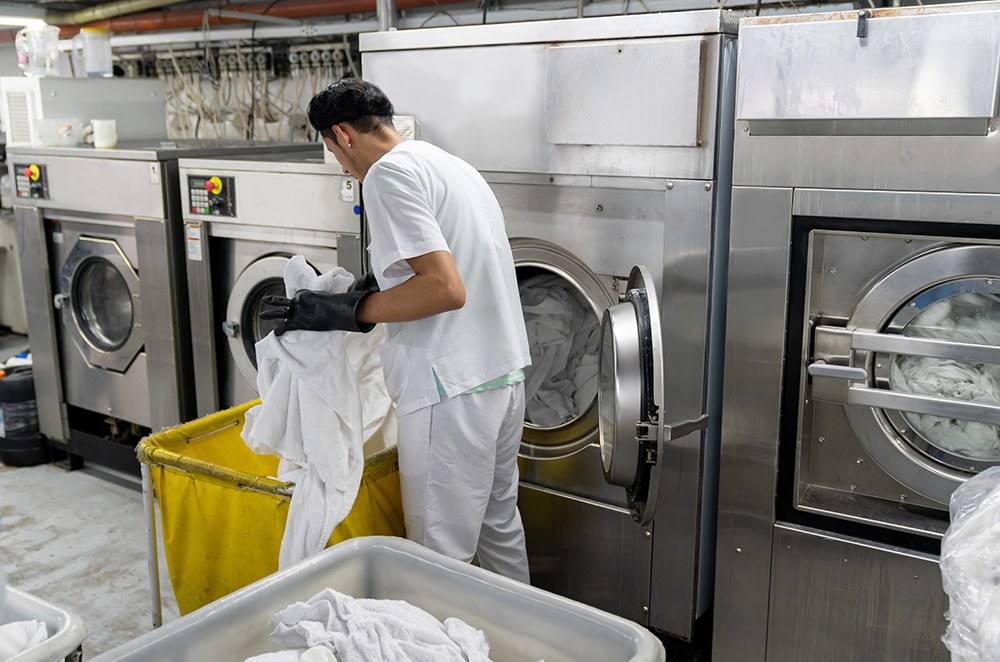 Mann nimmt Weißwäsche aus einer Großwaschmaschine in einer Wäscherei.