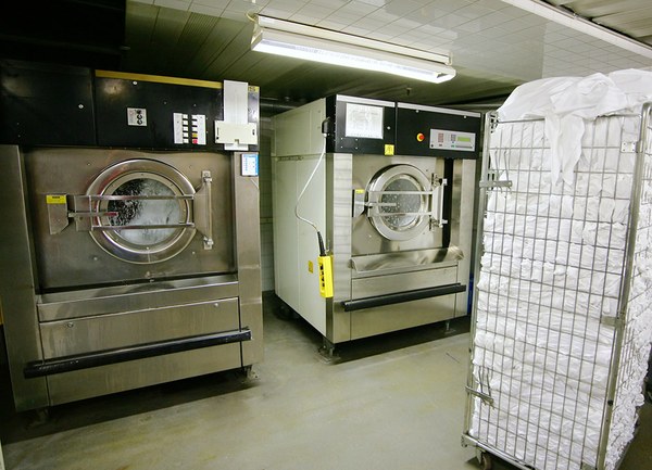 Zwei Großwaschmaschinen in einer Wäscherei in Betrieb, an der Seite ein Behälter mit Weißwäsche.