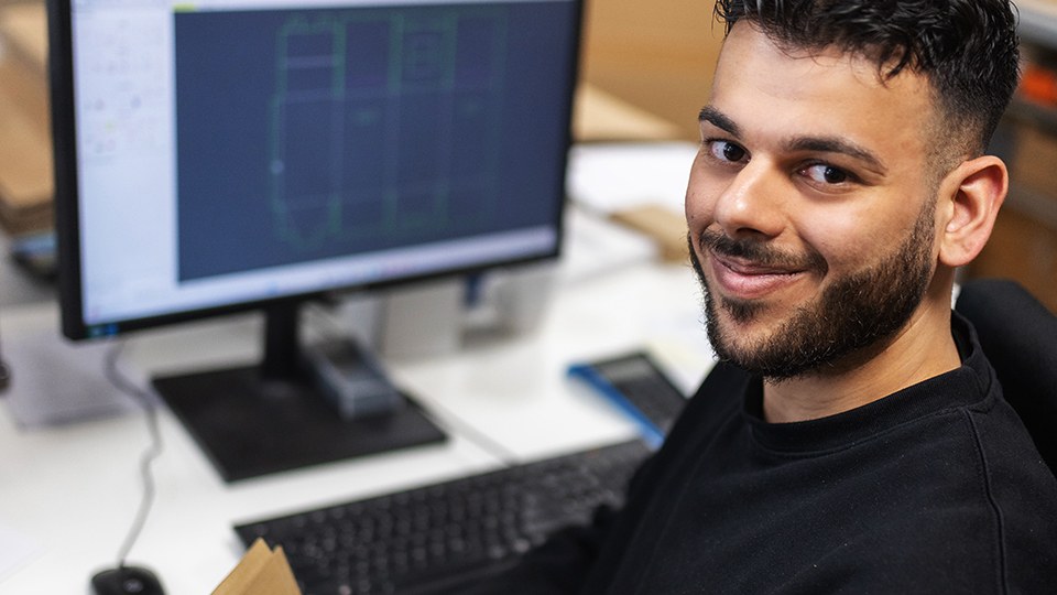 Sergen Sökmez an seinem Schreibtischarbeitsplatz; auf dem Monitor ist die Abwicklung einer Verpackung zu sehen; er hält ein Pappteil in den Händen und blickt in die Kamera